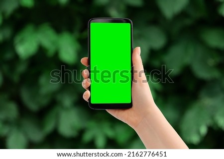 Hand with the phone upright against blurred greens. Smartphone with a green screen, copy space.