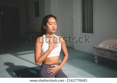 Asian woman practicing yoga in Diaphragmatic breath pose at spacious home. Concept of harmony and mental health. Young calm beautiful girl with closed eyes wearing sportswear on fitness mat Royalty-Free Stock Photo #2162778685