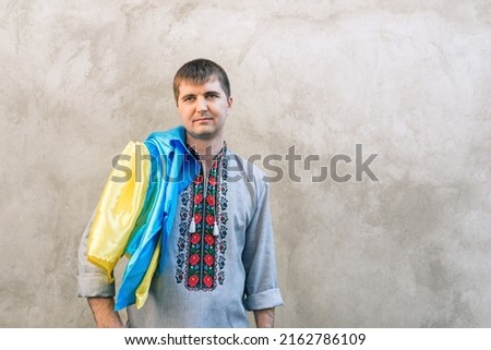 Portrait of a man holding the blue and yellow national flag of Ukraine on his shoulder and looking away. The guy in a traditional embroidered shirt on a background of a concrete wall. Copy space.