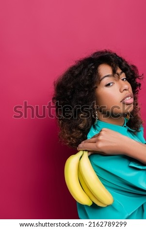 brunette and curly african american woman with fresh bananas looking at camera isolated on pink