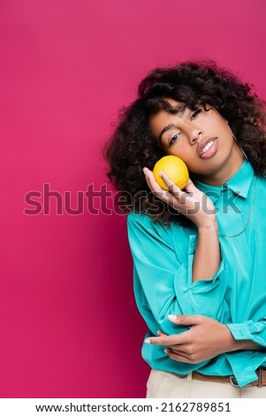 african american woman with wavy hair holding ripe orange isolated on pink