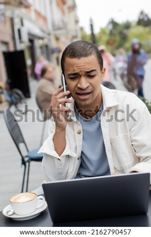 young african american freelancer talking on smartphone near laptop on cafe terrace