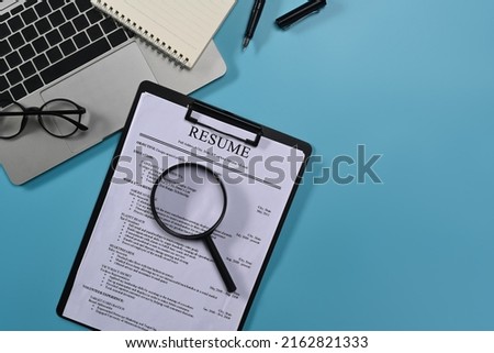 Top view laptop computer, magnifying glass and resumes applicants on blue background. Job search concept