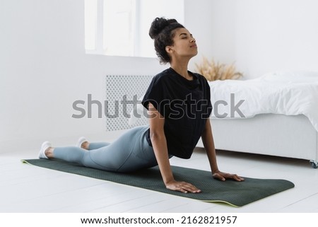 Side view of afro american female making breathing exercise, backbend and doing upward-facing dog pose or urdhva mukha svanasana. Woman at sport training at home, wearing in sportswear Royalty-Free Stock Photo #2162821957