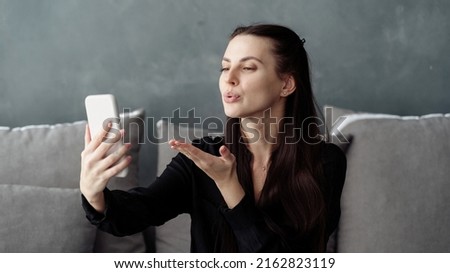 Side view of flirty young woman using online app on modern smartphone, talk on video call, send kiss. Female sitting on comfort couch in living room, making self portrait on front camera