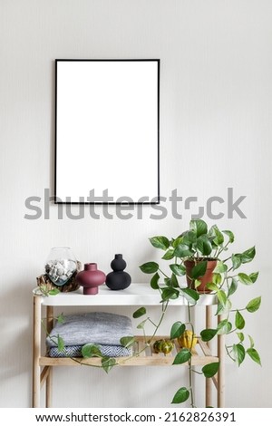 Picture frame with scenic ocean view hanging on wall over home decor and green potted plant. Vertical shot of vase with seashells standing on minimalistics white table in hygge apartment