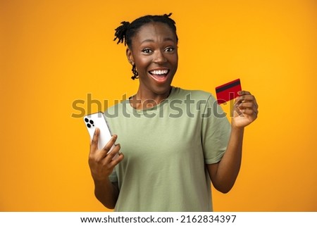 Smiling young afro girl holds smart phone and credit card in yellow studio
