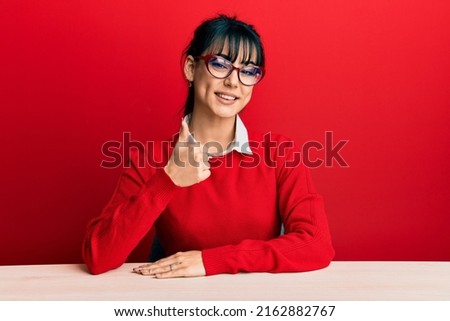 Young brunette woman with bangs wearing glasses sitting on the table doing happy thumbs up gesture with hand. approving expression looking at the camera showing success. 