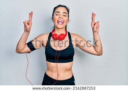 Young hispanic girl wearing gym clothes and using headphones gesturing finger crossed smiling with hope and eyes closed. luck and superstitious concept. 