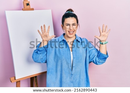 Young hispanic woman standing by painter easel stand showing and pointing up with fingers number ten while smiling confident and happy. 