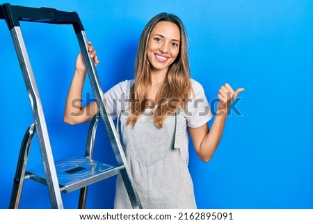 Beautiful hispanic woman standing by ladder pointing thumb up to the side smiling happy with open mouth 