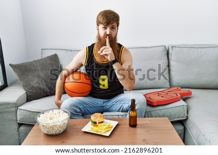 Caucasian man with long beard holding basketball ball cheering tv game asking to be quiet with finger on lips. silence and secret concept. 