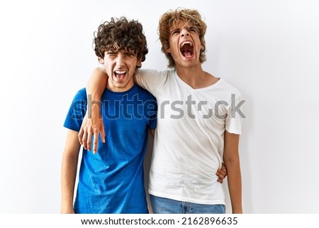 Young gay couple standing together over isolated background  screaming frustrated and furious, shouting with anger. rage and aggressive concept. 