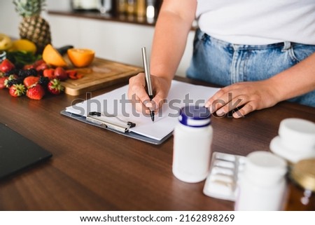 Closeup cropped photo of plump plus size woman counting calories, writing on clipboard healthy food, medicines, pills containers. Nutrition concept