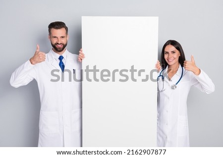 Portrait of two attractive cheerful medics holding big poster copy space ad showing thumbup isolated on grey color pastel background