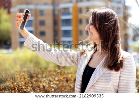 Young woman making selfie in beautiful garden in sunny day. Playful girl having fun with cellphone camera. Attractive woman smiling in smartphone camera in park