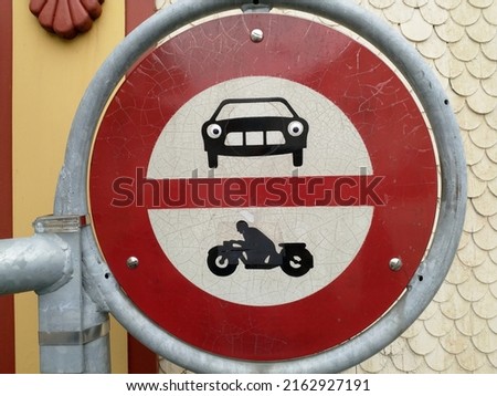 Street sign prohibiting trespassing for cars and motorcycles through. The sign is red and white. Someone put funny eyes on the cars lights.