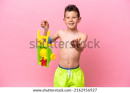 Little caucasian boy holding beach toys isolated on pink background inviting to come with hand. Happy that you came