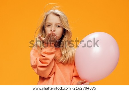  sweet girl, of school age, stands on an orange background in bright clothes, with a balloon in her hand and sends an air kiss. Horizontal studio photography with blank space for advertising mockup