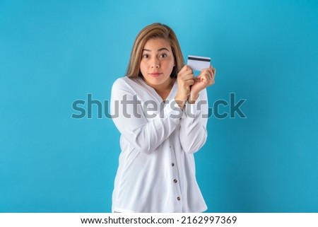 Beautiful attractive young business woman shocked while holding credit card, in white shirt and grey pants. Isolated over blue background.