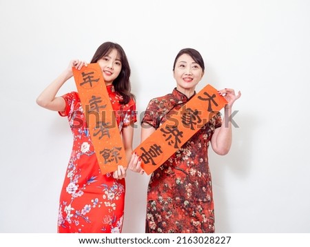 Asian women both wearing traditional cheongsam qipao dress hold Chinese festival couplet to celebrate new year with word meaning "May there be surplus year after year" on white background. Royalty-Free Stock Photo #2163028227