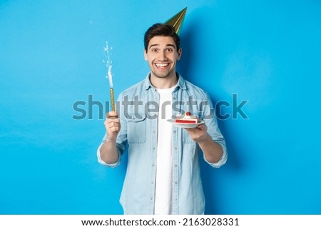 Happy young man celebrating birthday in party hat, holding b-day cake and smiling, standing over blue background