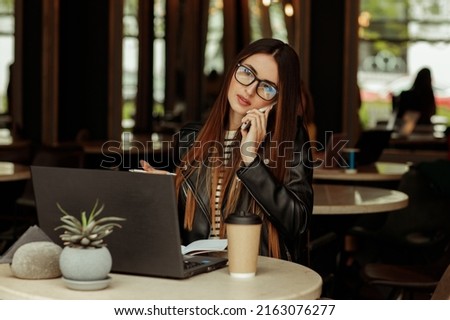 A young beautiful freelancer woman is working on a laptop and talking on the phone in a coffee shop