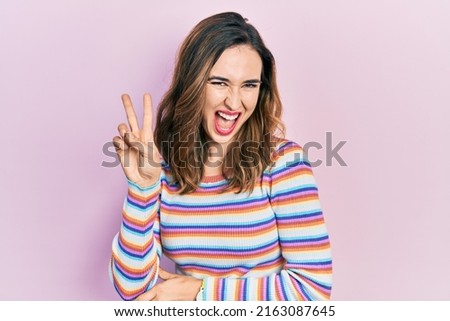 Young hispanic girl wearing casual clothes smiling with happy face winking at the camera doing victory sign with fingers. number two. 