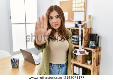 Young brunette woman holding solar windmill for renewable electricity at the office with open hand doing stop sign with serious and confident expression, defense gesture 