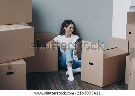 Happy spanish woman getting message from shipping service. Lady is packing boxes for the relocation. Girl is chatting on mobile phone and sitting on the floor. Easy moving concept. Royalty-Free Stock Photo #2163093411
