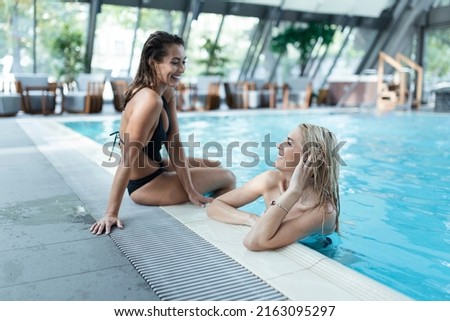 Two girlfriends in swimwear sitting on the poolside, enjoying summer vacation on the swimming pool intdoors