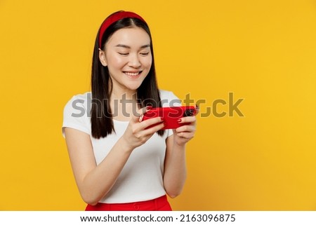 Fun young woman of Asian ethnicity she 20s wears white t-shirt using play racing app on mobile cell phone hold gadget smartphone for pc video games isolated on plain yellow background studio portrait