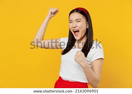 Happy fun excited jubilant young woman of Asian ethnicity 20s years old wears white t-shirt doing winner gesture celebrate clenching fists say yes isolated on plain yellow background studio portrait Royalty-Free Stock Photo #2163096901