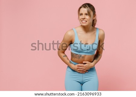 Young strong sporty athletic fitness trainer instructor woman wear blue tracksuit spend time in home gym put hand on belly suffer look aside isolated on plain pink background. Workout sport concept Royalty-Free Stock Photo #2163096933