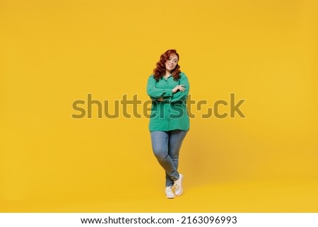 Full size body length bright happy vivid fancy young ginger chubby overweight woman 20s wears green shirt looking camera smiling hold hands crossed isolated on plain yellow background studio portrait