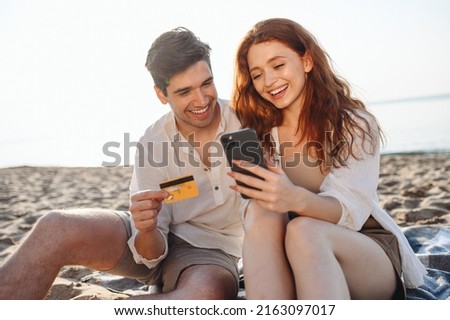 Young couple two man woman 20s in white clothes sit on sand use mobile phone credit card shopping online order delivery book tour rest together at sunrise over sea beach outdoor seaside in summer day