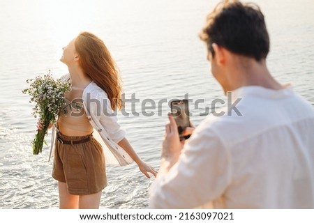 Tender young couple family man woman in white clothes hold flowers bouquet take photo on mobile cell phone rest together at sunrise over sea beach ocean outdoor seaside in summer day sunset evening.