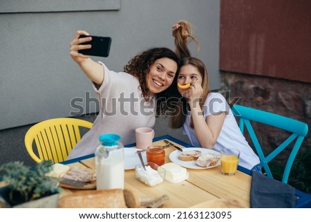 young mother mother and her teenager daughter having breakfast in home backyard . taking selfie with smartphne and making funny faces