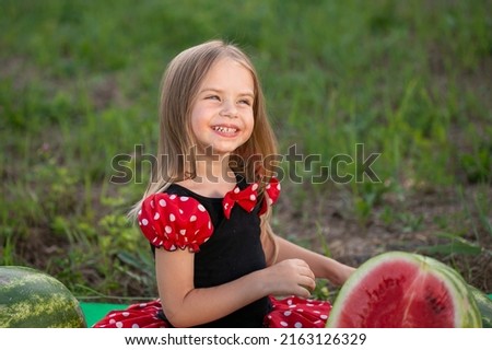 Beautiful little blonde girl, has happy big eyes, fun smile, eating watermelon in nature garden. Child portrait. Lifestyle concept. Summer time. Fashion kid style. Amazing face.
