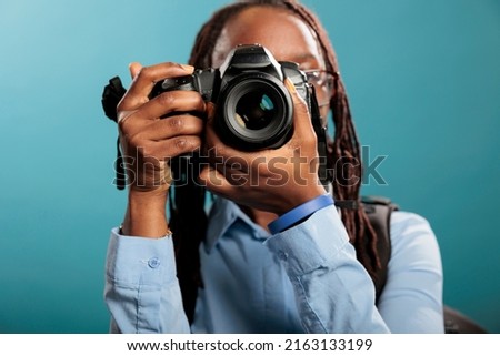 Studio shot of young adult woman with DSLR camera shooting photos while standing on blue background. Professional photographer with photography modern device taking pictures.