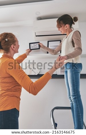 Mother and daughter on the ladder hanging pictures and photos on the shelf and wall at home.