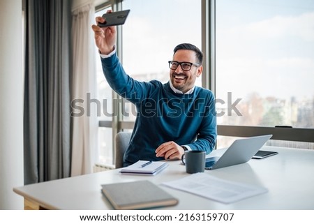 Excited young businessman taking selfie on smart phone. Happy professional in smart casuals is sitting at desk. He is working by window in office.