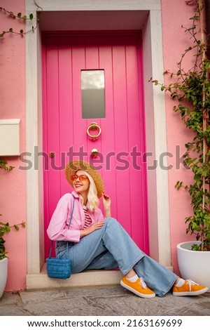 Happy smiling traveler woman posing in street, near pink color house. Model wearing trendy summer outfit with sunglasses, straw hat, shirt, wide jeans, orange sneakers, shoulder blue bag Royalty-Free Stock Photo #2163169699