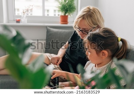 Granny and preschool girl using tablet together at home on the sofa, family togetherness time 