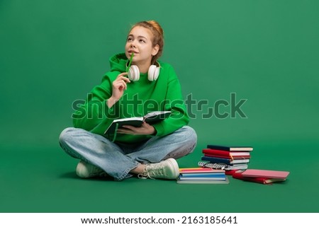 Portrait of young girl, student in casual cloth sitting on floor with thoughtful expression, studying isolated over green studio background. Concept of education, studying, homework, youth, lifestyle Royalty-Free Stock Photo #2163185641