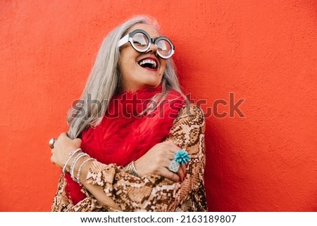 Cheerful senior woman laughing happily while standing against a red background. Carefree elderly woman feeling youthful and vibrant in colourful clothing. Mature woman enjoying life after retirement. Royalty-Free Stock Photo #2163189807