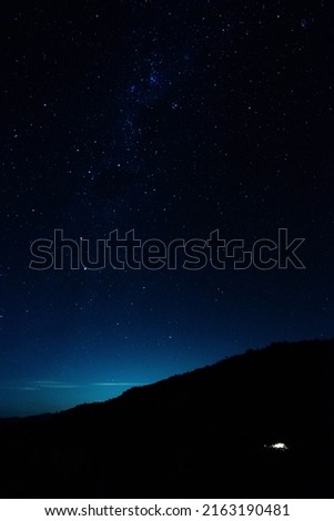 night photo with the sky a house on a mountain and the light of a city from afar, black background with stars

