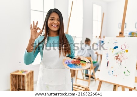 Young latin woman standing at art studio doing ok sign with fingers, smiling friendly gesturing excellent symbol 