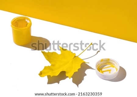 Minimal composition with yellow maple leaf and jar of yellow paint on white and yellow background. Arrives autumn concept. Fall of the leaves, change of seasons concept.