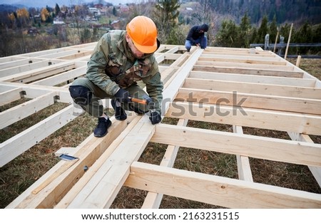 Man worker building wooden frame house on pile foundation. Carpenter hammering nail into wooden board, using hammer. Carpentry concept. Royalty-Free Stock Photo #2163231515